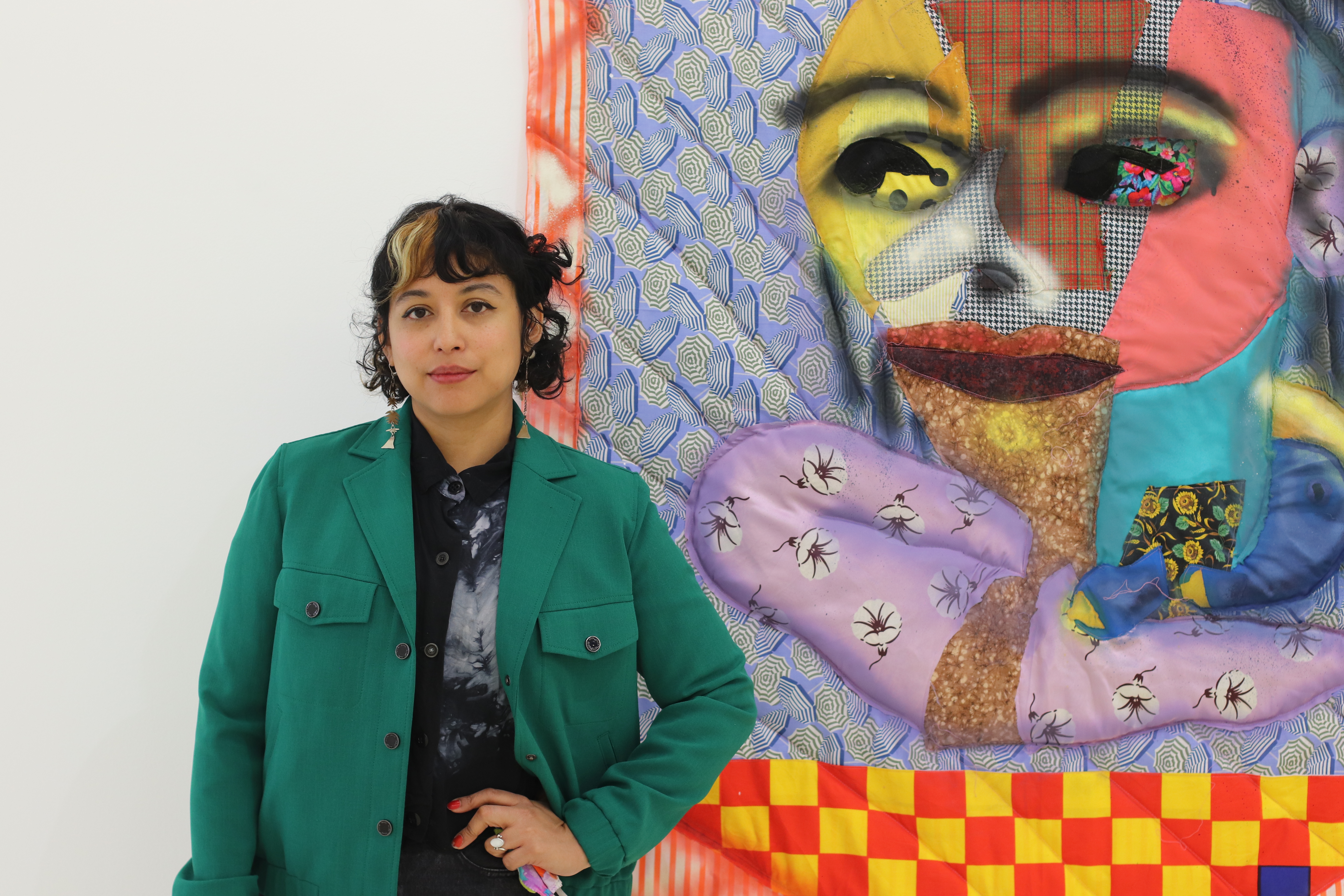 Maria Guzmán Capron standing in front of a colorful quilted artwork depicting a face.