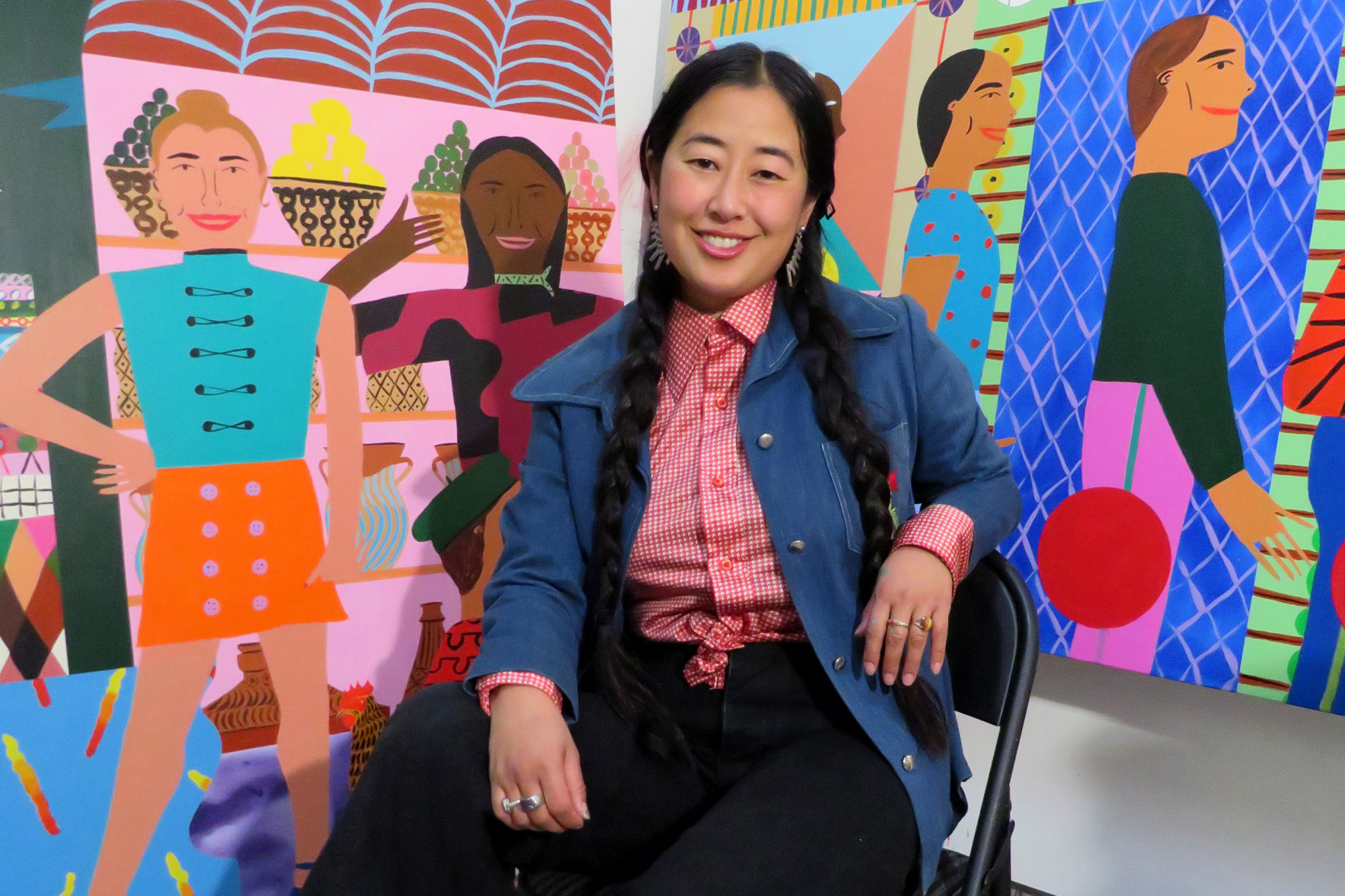Chelsea Ryoko Wong sitting in a chair in front of two colorful paintings depicting people.