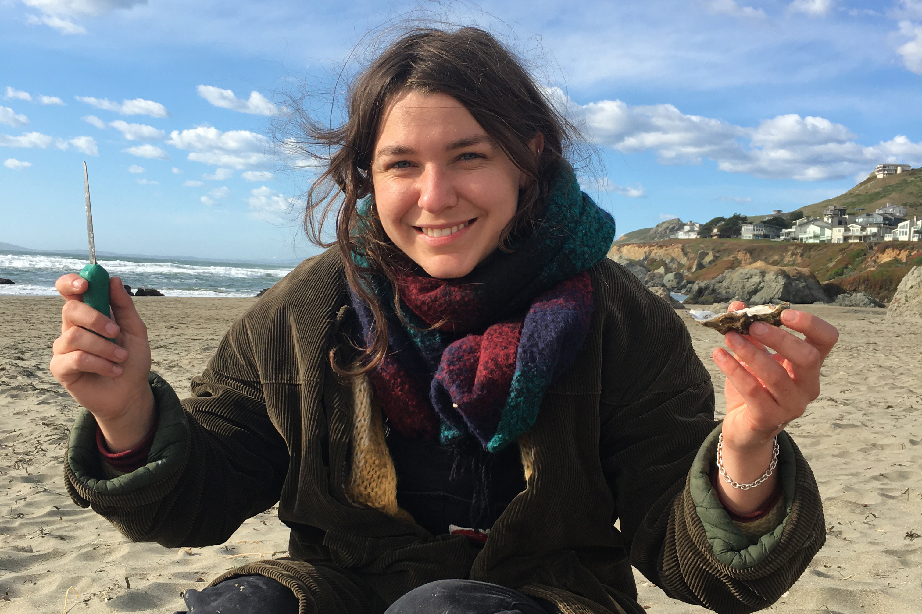 Paige Valentine sitting on a beach holding up an oyster and an oyster shucking tool.