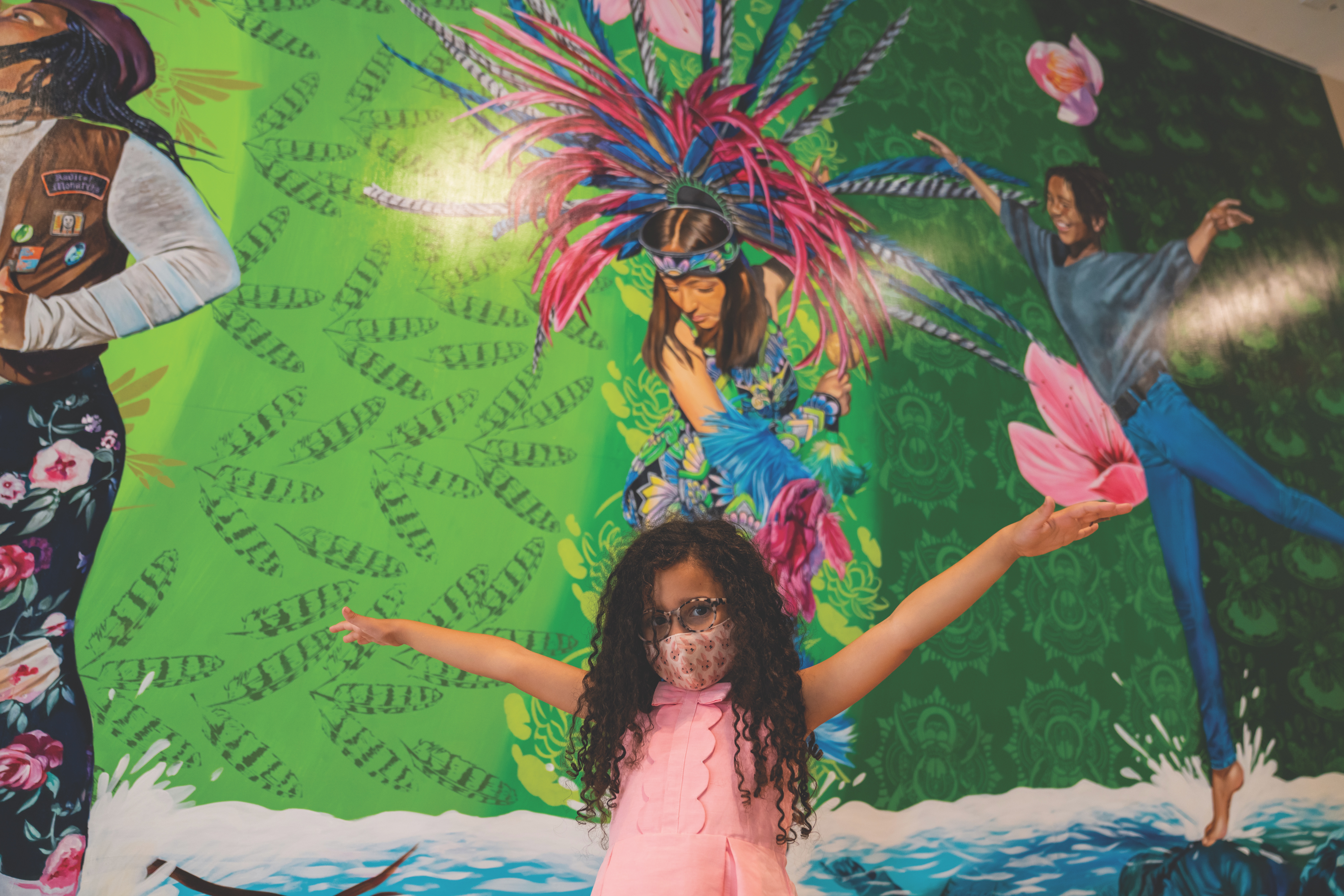 A young girl poses with her arms in the air in front of Twin Walls Mural Company's mural "Our Ancestors Wildest Dreams" depicting various women and girls dancing.